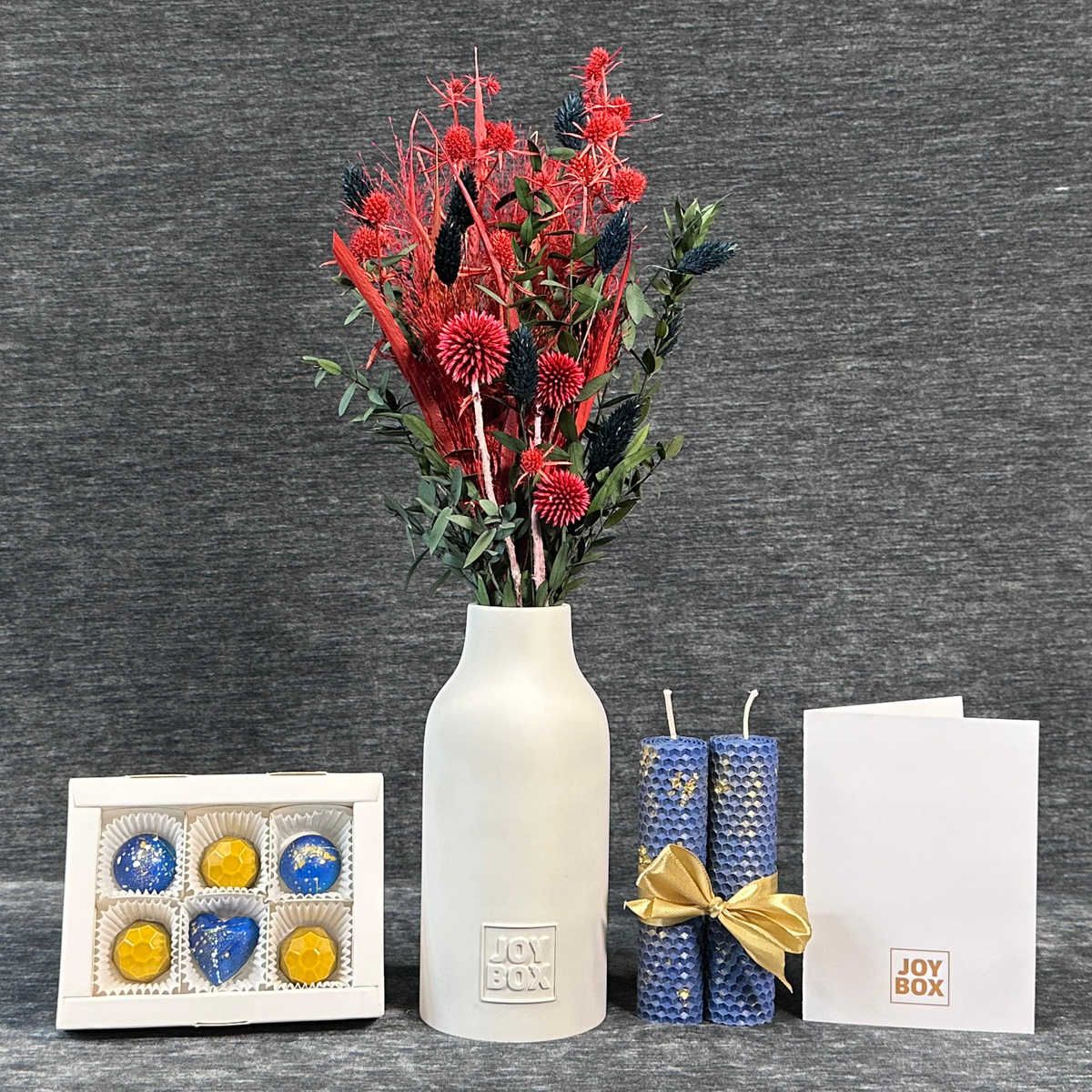 Подарунковий набір «Рубіновий зорепад» JOY BOX: букет сухоцвітів у білій гіпсовій вазі, сині вощинні свічки, коробка синьо-золотих цукерок і біла листівка на сірому фоні